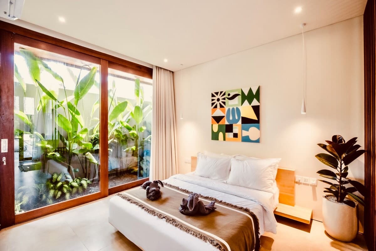 Queen bed dressed in white linen beneath a geometric print, sliding doors open to a leafy garden outlook