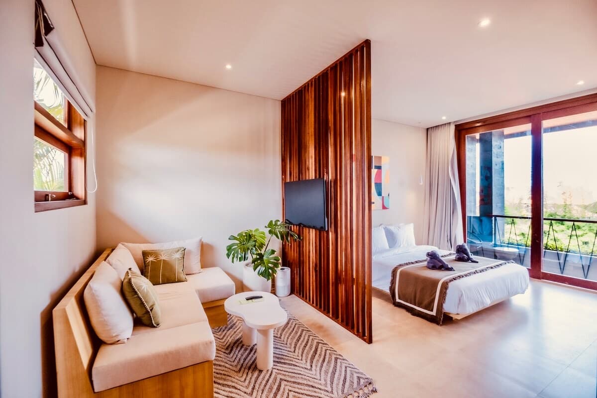 Open-plan studio with a linen sofa, wall-mounted TV and queen bed, balcony doors open to greenery beyond